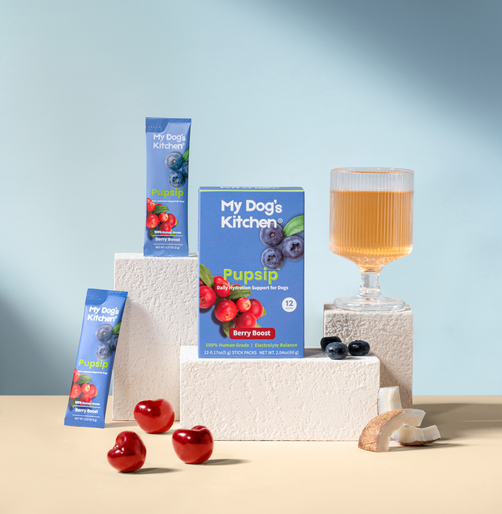 Stylized product still-life featuring PupSip Berry Boost: a box and individual Berry Boost stick packs displayed on textured white blocks, alongside a glass of amber-colored drink and scattered blueberries, cranberries, and coconut pieces on a light blue backdrop—highlighting the natural ingredients and premium hydration experience.